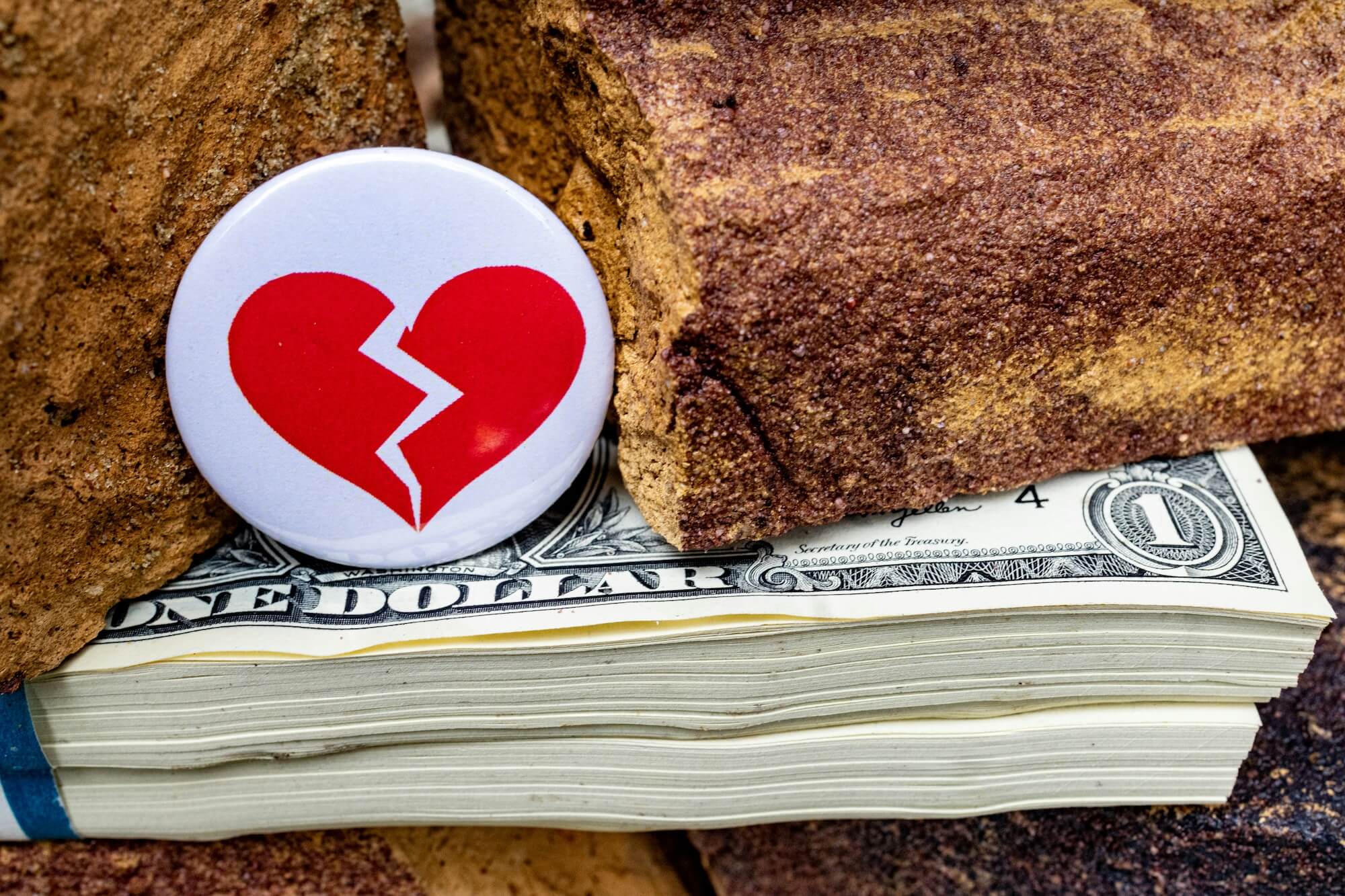 A broken heart symbol pin sits atop of a stack of U.S. currency.